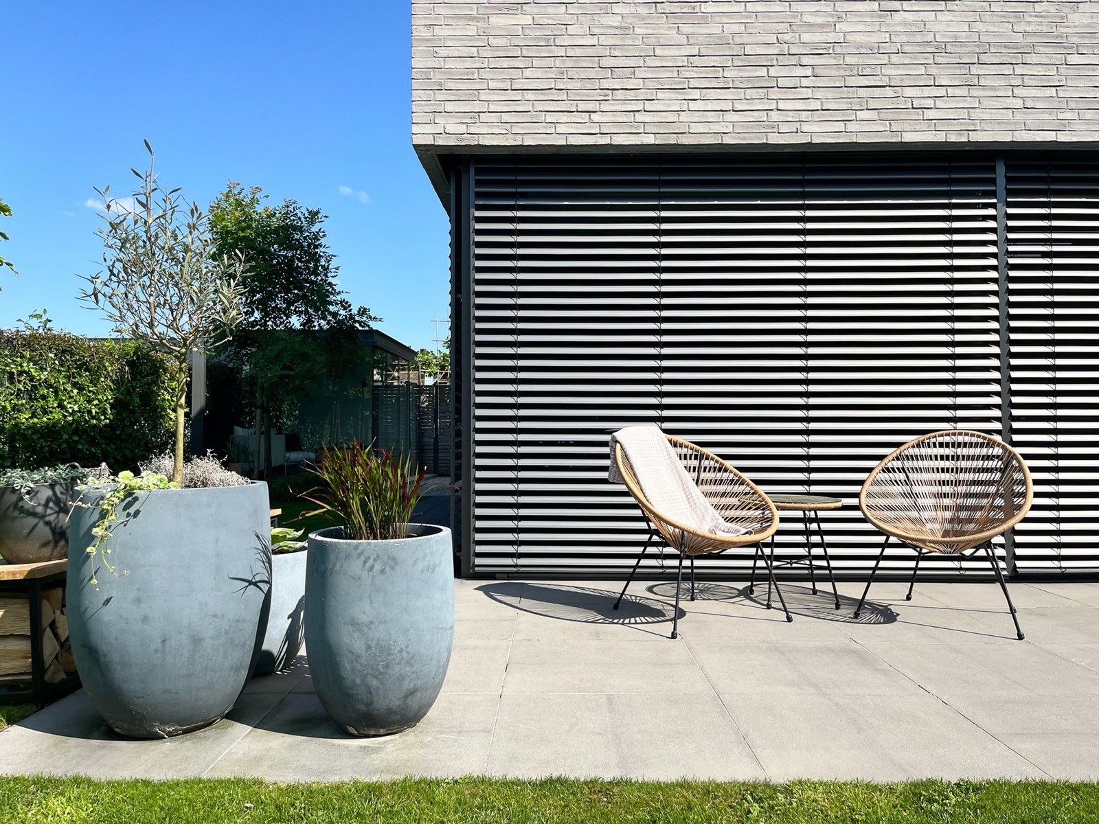 Sonnige Gartenterrasse mit Rattan-Sitzgruppe, Betonpflanzgefäßen und dunkler Lamellen-Verschattung.
