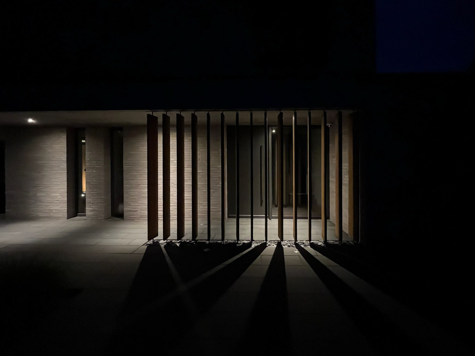Hauseingang bei Nacht — vertikale Holzlamellen werfen lange Schatten, warmes Licht im Inneren.